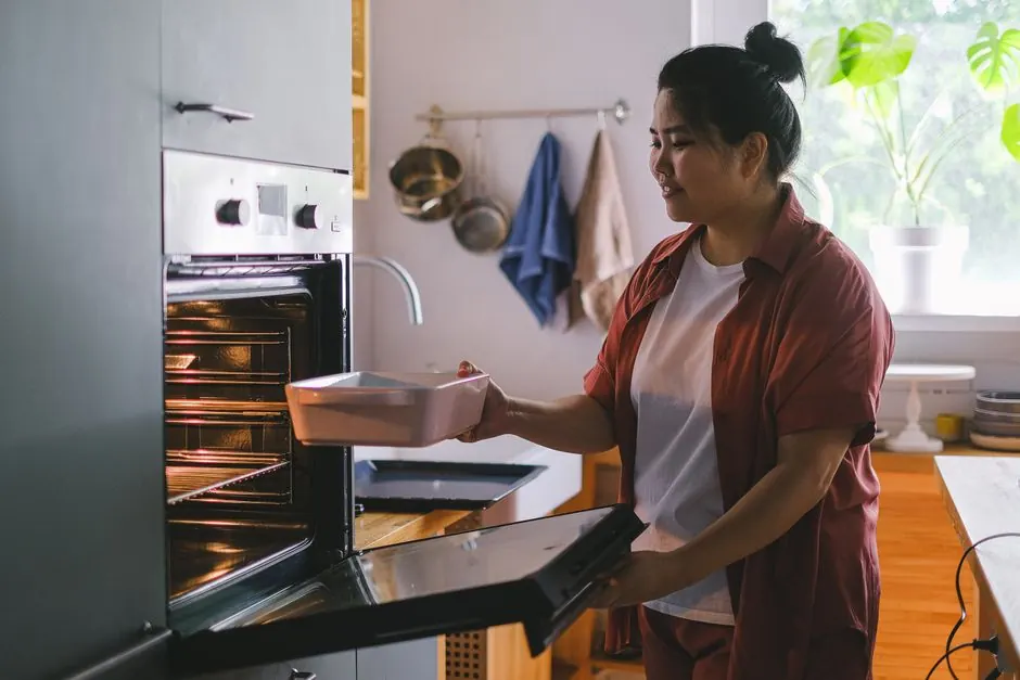 A woman prepares to bake by placing a dish in the oven in a sunny kitchen.